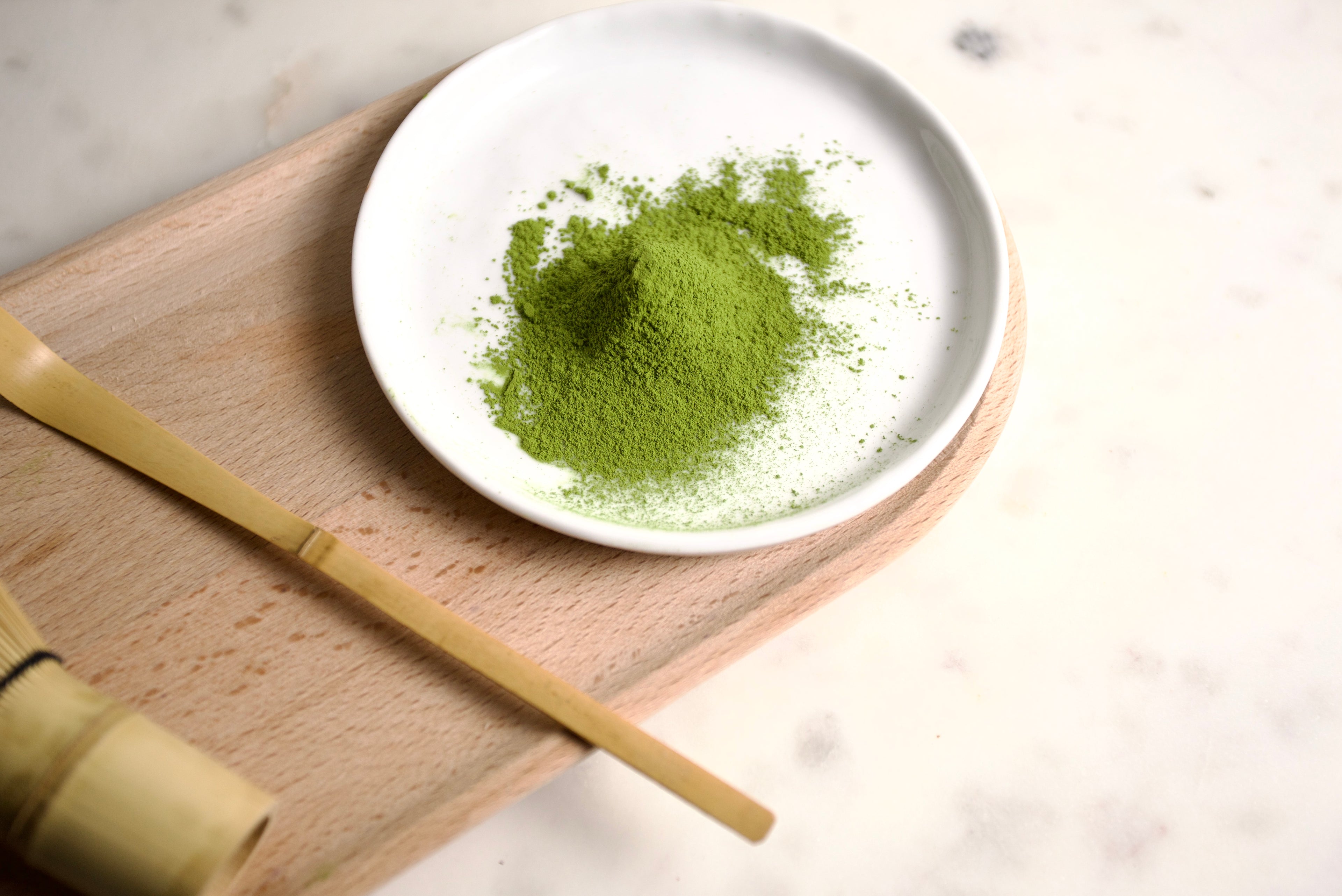 A small mound of bright green matcha powder sits in a white ceramic dish on a light wooden board, with a bamboo whisk visible in the lower left on a pale marble surface.